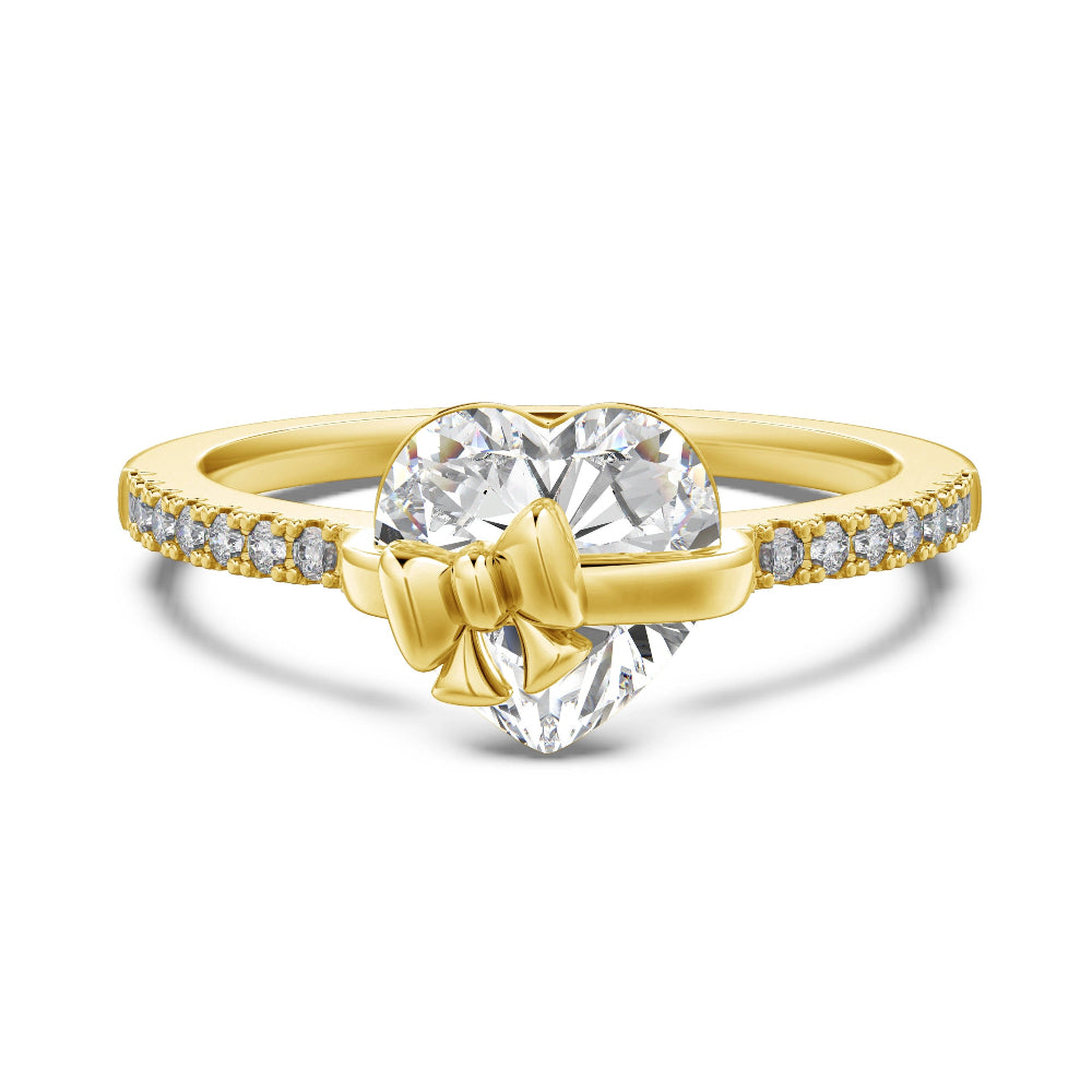 A yellow gold ring featuring a heart-shaped cubic zirconia in the center, adorned with a small gold bow in front. The band is paved with small round clear cubic zirconia s on either side.